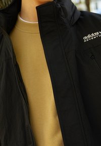 Svart vattentålig jacka med hög krage, fickor med dragkedja och "adidas Adventure"-logotyp. Under jackan bärs en ljusbrun sweatshirt.