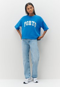 Zils pārmērīga izmēra džemperis ar uzrakstu "PORTO" baltā krāsā, kombinēts ar gaiši zilām džinsa biksēm un baltām sporta kurpēm. Gluda auduma un brīvs piegriezums.