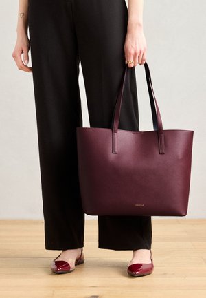 Bordeaux tote bag med tekstureret overflade og matchende hanke, forsynet med et guld logo, holdt af en person iført sorte brede bukser.