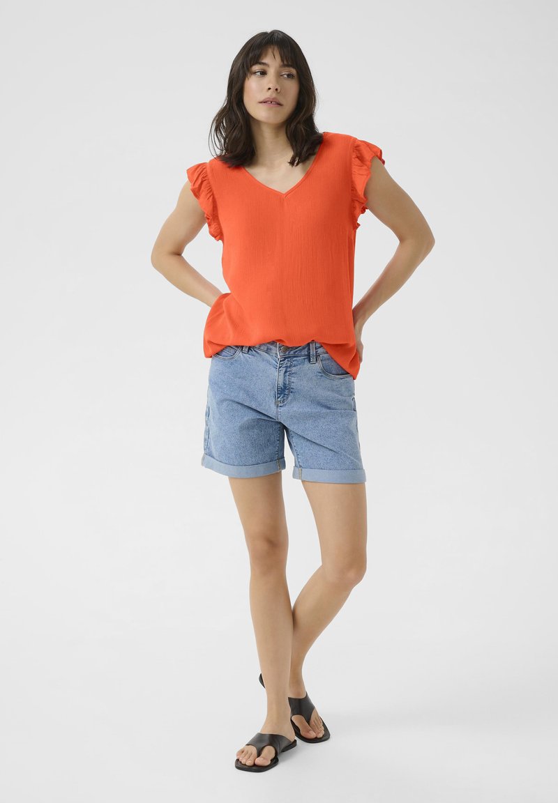 Haut orange vif à manches courtes avec des bords volants ; associé à un short en denim bleu clair avec des poignets retroussés et des sandales plates noires.