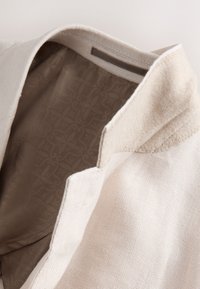 Vêtement en lin beige avec un col texturé doux et une doublure intérieure à motif. Présente des coutures nettes et un design minimaliste.