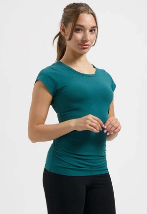 Junge Frau mit zurückgebundenem braunem Haar, die ein enganliegendes, kurzärmeliges Shirt in Petrol und schwarze Hose trägt, steht vor einem einfarbigen Hintergrund.