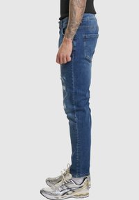 Jean en denim bleu slim fit avec des détails usés, design à cinq poches et couture visible. Porté avec des chaussures de course argentées.