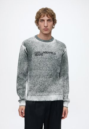 Mann mit hellbraunem lockigem Haar, der einen ausgewaschenen grauen Pullover mit dem Schriftzug „KARL LAGERFELD JEANS“ und schwarze Hose vor weißem Hintergrund trägt.