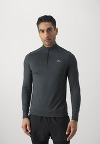 Tmavomodrý dlhý rukáv športový top na zips s elastickým materiálom, s malým zeleným logom na hrudi a priliehavým strihom.