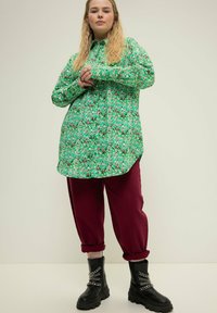 Camicia verde a fantasia con colletto, abbinata a pantaloni tapered bordeaux e stivali neri stringati con dettagli in catena.