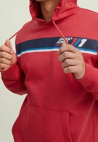 Persona vistiendo una sudadera roja con una franja negra, azul y blanca en el pecho y sosteniendo los cordones de la sudadera.