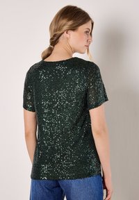 Model wearing a short-sleeve, dark green sequined top, facing away, with wavy hair held back, and blue jeans in a simple background.