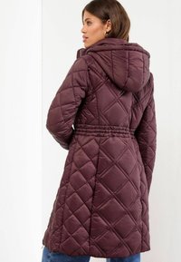 Vrouw in een lange, geplooide bordeauxrode jas met capuchon, gezien vanuit de rug, staand tegen een witte achtergrond.