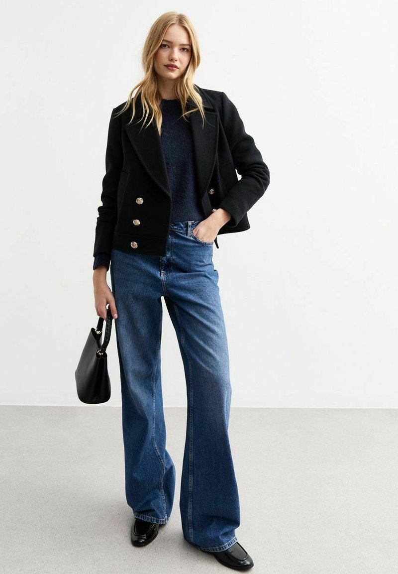 Svart dubbelknäppt jacka med guldknappar, tillsammans med vida blå denimjeans. Svart handväska, svart sko med spetsig tå och mörkt överdel.