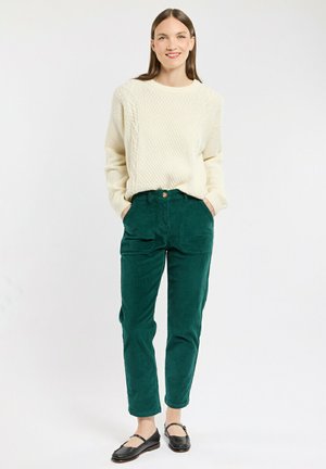 Un pull en tricot torsadé crème, associé à un pantalon en velours côtelé vert foncé avec fermeture à bouton, coupe droite et poches latérales. Des ballerines noires.