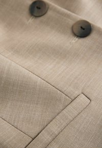 Blazer beige doppiopetto con due bottoni scuri, tessuto testurizzato e un elegante dettaglio della tasca con cuciture a contrasto.