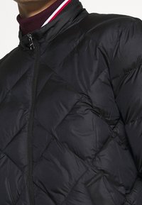 Giacca a vento nera con design a rombi, colletto alto e chiusura con zip. Accenti a coste in rosso e bianco vicino al collo.