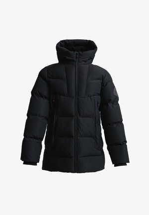 Jugendlicher mit lockigem Haar, der eine schwarze Steppjacke mit Kapuze und einem Canada-Logo-Patch am Oberarm trägt, steht mit den Händen in den Taschen.
