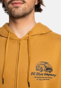 Geel ritsvest gemaakt van zacht materiaal met een autografiek en de tekst "DC Shoe Company" op de borst. Voorzien van een verstelbare capuchon en een metalen rits.