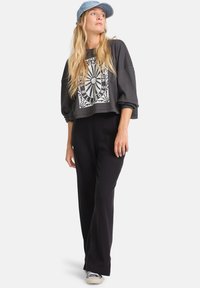 Grauer verkürzter Sweatshirt mit Grafikdesign, kombiniert mit schwarzen, weit geschnittenen Hosen. Eine blaue Denim-Cap und hellfarbige Sneakers runden den Look ab.