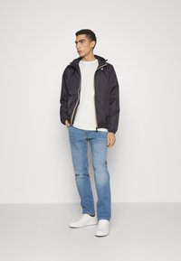 Jachetă neagră impermeabilă cu manșete elastice, detalii cu fermoar galben, purtată peste un tricou alb și blugi, completată cu adidași albi.