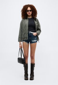 Γυναίκα με σγουρά μαλλιά που φοράει μαύρα γυαλιά ηλίου, πράσινο μπουφάν, μαύρο τοπ, σκισμένο τζιν σορτς, μαύρες μπότες και κρατάει μια μαύρη τσάντα χειρός.