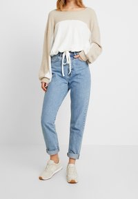 Pull en tissu côtelé beige clair et blanc avec une taille à cordon, associé à un jean bleu clair retroussé et des baskets beiges.