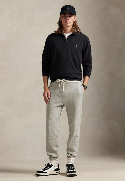 Junger Mann, der einen schwarzen Pullover mit Viertel-Reißverschluss, graue Jogginghose, schwarze Kappe und schwarz-weiße Sneakers trägt und vor einem schlichten Hintergrund steht.