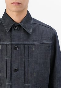 Mørk denimjakke med spidse kraver, frontknaplukning og stikninger. Stoffet har en tekstureret, let slidt udseende.