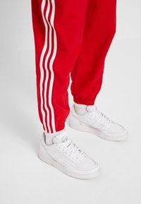 Röda träningsbyxor med vita sidostyklinjer och elastiska muddar, tillsammans med vita sneakers med en slät läderstruktur och perforeringar.