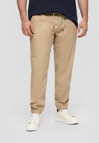 Beige lichtgewicht joggers met elastische boorden en een verstelbare trekkoord tailleband, gedragen met witte sneakers. Soepele textuur, minimalistisch ontwerp.
