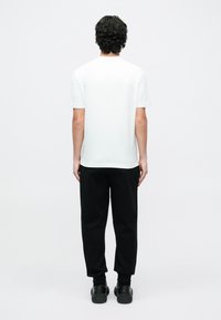 Muž s kučeravými vlasmi, ktorý má na sebe jednoduché biele tričko s krátkym rukávom, čierne teplákové nohavice a čierne tenisky, stojaci chrbtom čelom na bielom pozadí.