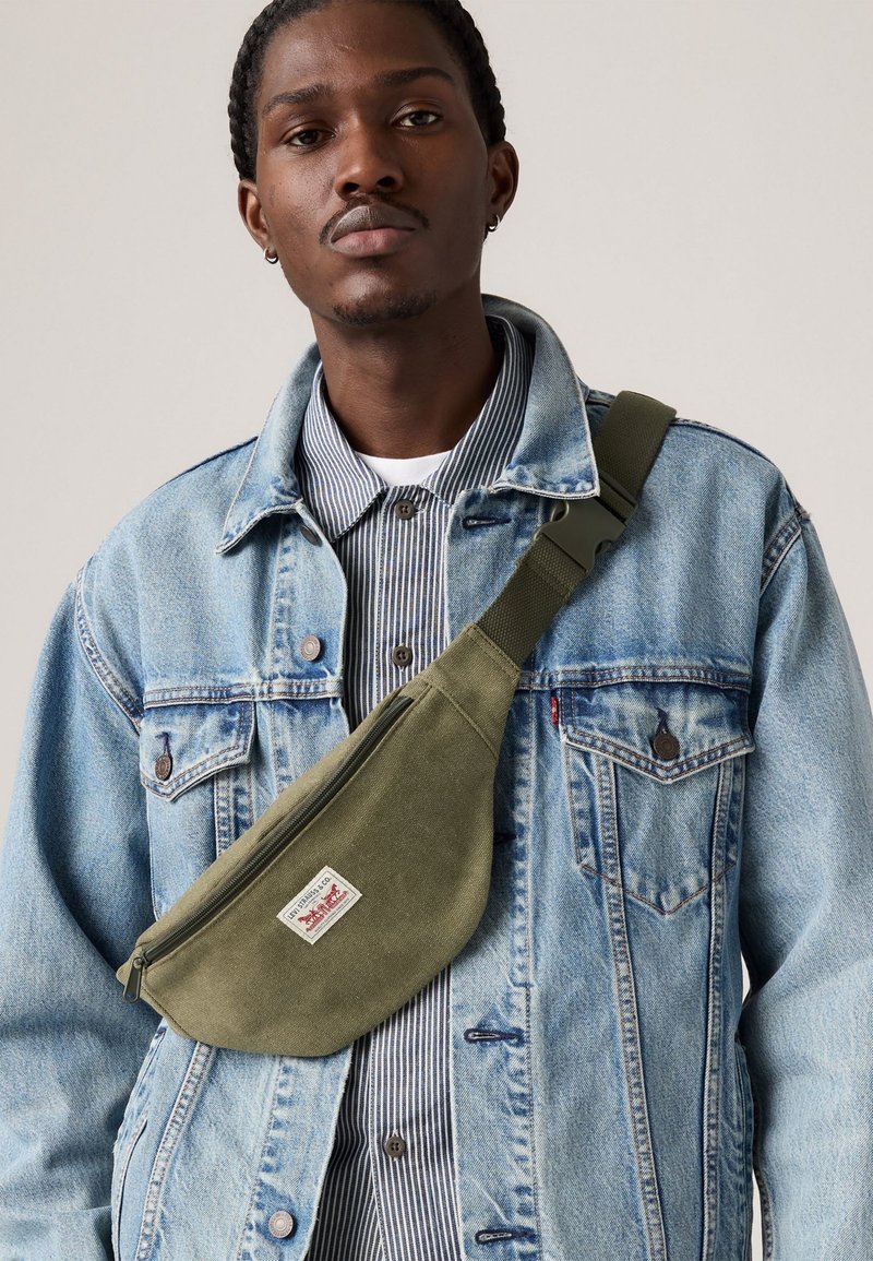 Man in een blauwe spijkerjack, gestreept shirt en een olijfgroene schoudertas met het Levi's logo aan de voorkant, tegen een neutrale achtergrond.