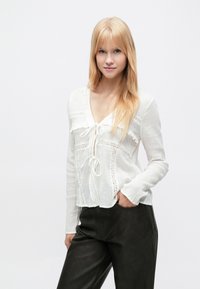 White long-sleeve blouse with a V-neck and tie closure, featuring lace trim and textured fabric. Paired with dark, fitted pants.