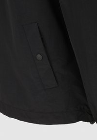 Détail de la veste noire avec un tissu lisse et léger, une poche latérale avec fermeture à bouton et un ourlet plat.