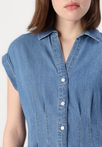 Camicia di jeans blu con maniche corte arrotolate, chiusura frontale con bottoni e cuciture verticali. Tessuto morbido con una vestibilità strutturata.
