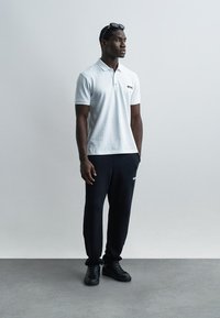 Άνδρας που φοράει λευκό πόλο πουκάμισο, μαύρο παντελόνι, μαύρα αθλητικά παπούτσια και γυαλιά ηλίου τοποθετημένα στο κεφάλι, στέκεται μπροστά σε απλό γκρι φόντο.