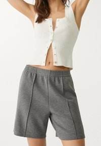 Woman in white sleeveless buttoned top and gray elastic waist shorts, arms raised against plain light background.