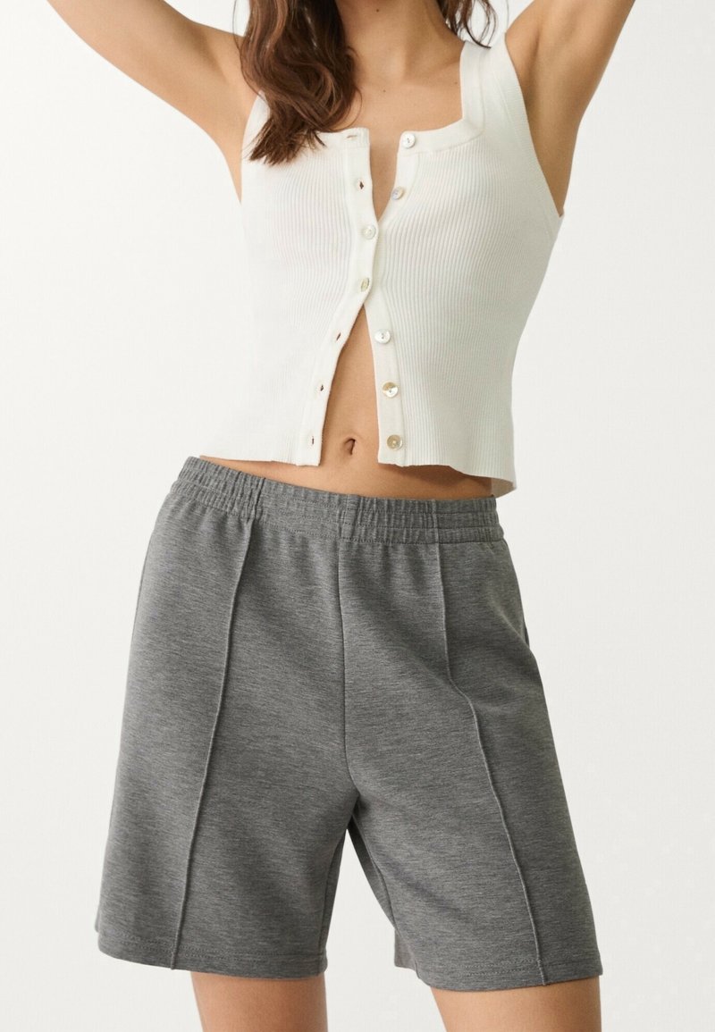 Woman in white sleeveless buttoned top and gray elastic waist shorts, arms raised against plain light background.