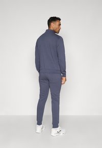 Námornícky modrý športový komplet z mäkkej látky, s golierom na zips a elastickými manžetami, v kombinácii s bielymi športovými topánkami. Model zobrazený zozadu.