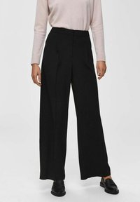 Zwarte wijde pantalon met een hoge taille en voorplooien, gemaakt van een gladde stof. Gepaard met een lichtroze blouse met lange mouwen en zwarte schoenen.
