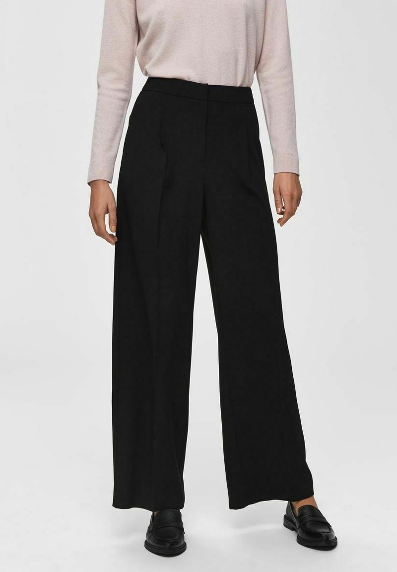 Zwarte wijde pantalon met een hoge taille en voorplooien, gemaakt van een gladde stof. Gepaard met een lichtroze blouse met lange mouwen en zwarte schoenen.