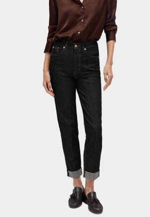 Kvinde iført mørkebrun satin skjorte med knapper, højtaljede sorte bukser med manchetter og sorte loafers med frynser mod en ensfarvet lys baggrund.
