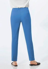 Pantalons bleu à taille élastique avec une coupe slim, présentant une texture lisse et des lignes épurées, se terminant juste au-dessus de la cheville. Portés avec des pantoufles noires plates.