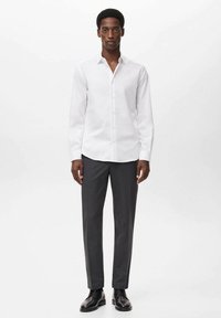 Camicia bianca a maniche lunghe con bottoni frontali, abbinata a pantaloni grigio scuro e scarpe nere. Tessuto morbido, colletto classico, vestibilità sartoriale.