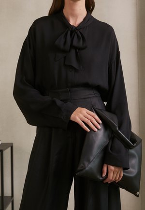 Femme portant un chemisier noir à col lavallière et un pantalon à jambes larges, tenant un grand sac pochette en cuir noir avec les deux mains.