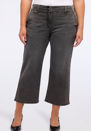 Jean noir délavé taille mi-haute, court et large, porté avec des talons noirs à bout pointu et un haut blanc, montré de la taille aux pieds.