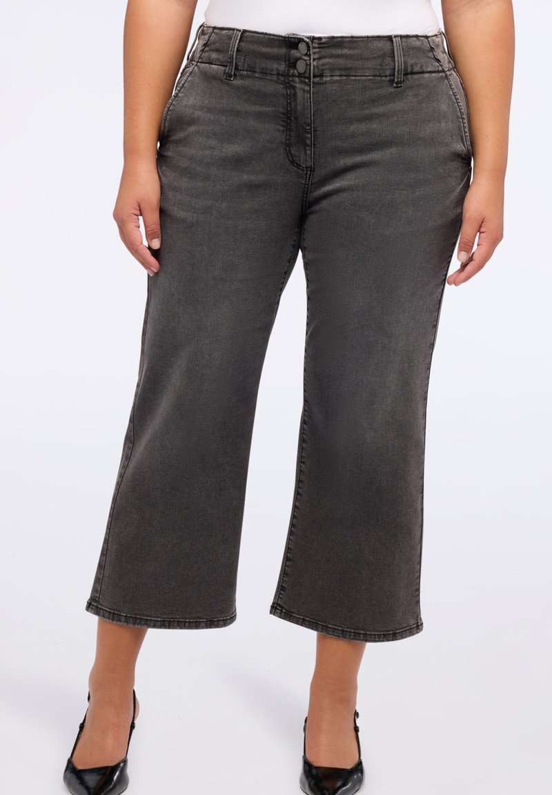 Jean noir délavé taille mi-haute, court et large, porté avec des talons noirs à bout pointu et un haut blanc, montré de la taille aux pieds.