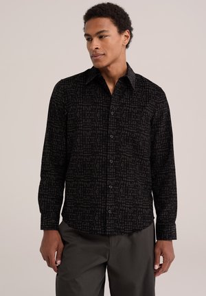 Jeune homme portant une chemise noire à motifs à manches longues et un pantalon foncé, regardant légèrement sur le côté sur un fond uni.