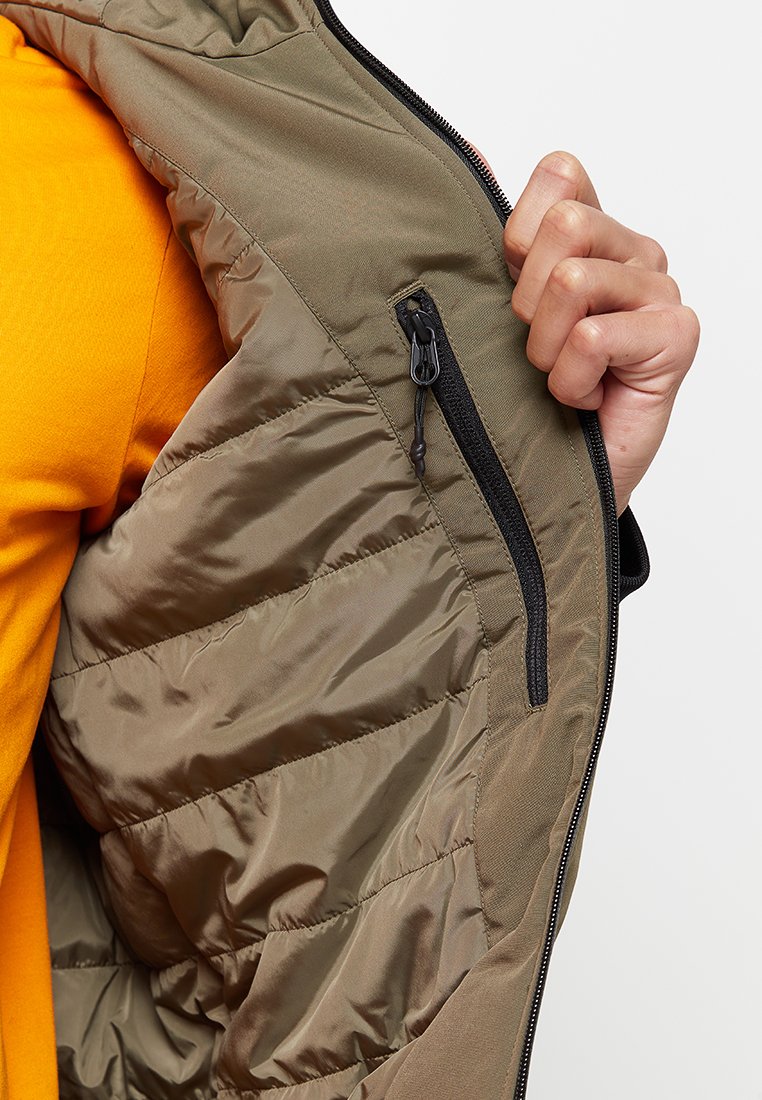 Gilet imbottito verde oliva con tasca laterale con zip, caratterizzato da tessuto trapuntato e una superficie liscia. Dettagli e fodera nera visibili.