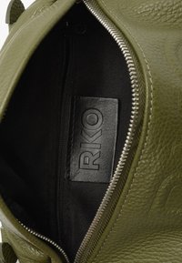 Intérieur d'un sac en cuir vert olive avec une doublure en tissu noir, comprenant une poche zippée et une étiquette RKO. Surface texturée et fermeture éclair métallique visibles.