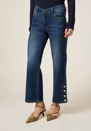 Donna che indossa jeans a flare cropped blu scuro con bottoni dorati vicino all'orlo, abbinati a scarpe con tacco a spillo a punta con stampa pitone.