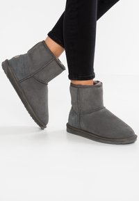 Stivaletti grigi alla caviglia in suede, con una morbida fodera interna, dettagli cuciti e suole in gomma piane, indossati con jeans neri.
