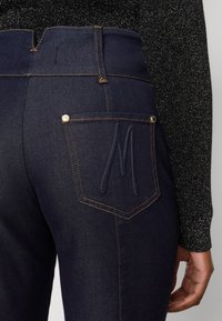 Mörkblå högmidjade jeans med synlig sömnad, en bakficka med en upphöjd 'M'-design och en metallknapp som accent.